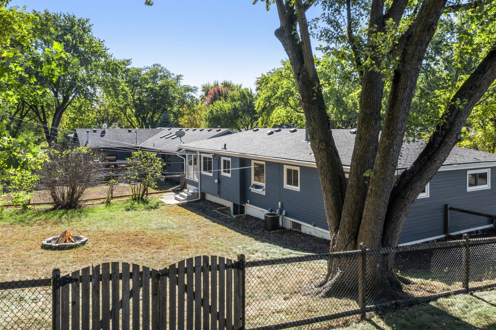 A fenced backyard with a large tree, a fire pit, and a gray single-story house with white trim, set among green trees under a clear blue sky.