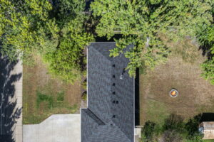 Aerial view of a house with a gray shingle roof, driveway on the left, green trees partially shading the yard, and a fire pit with chairs on the right side of the property.