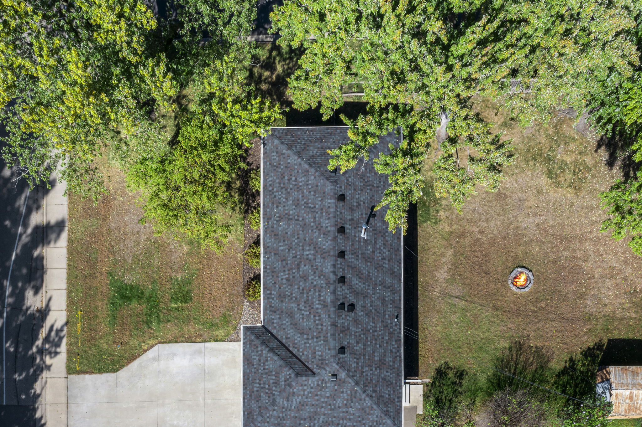 Aerial view of a house with a gray shingle roof, driveway on the left, green trees partially shading the yard, and a fire pit with chairs on the right side of the property.