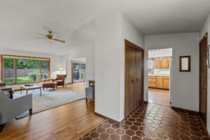 View from the entryway of 9333 Colorado Circle, a home for sale by Beekeeper Realty, featuring a tiled floor, wooden closet doors, and a hallway leading to a bright living room with large windows, light furniture, and a kitchen with wooden cabinets.