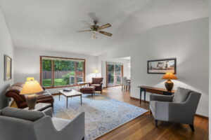 A bright living room at 9333 Colorado Circle features wood floors, large windows, a ceiling fan, leather sofa, armchairs, coffee table, area rug, lamps, and a view of the fenced green yard—an inviting Minnesota home for sale.