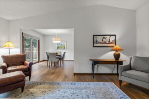 A cozy living room at 9333 Colorado Circle, Bloomington, features a leather armchair, gray chair, and area rug leading to a dining area. Soft lighting and wall art add warmth, while large windows frame Minnesota’s lush outdoor scenery.