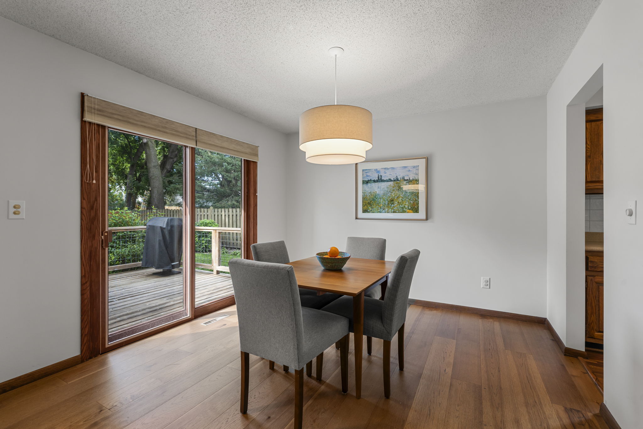 A dining room with a wooden table and four gray chairs, a large round ceiling light, and a sliding glass door leading to a deck with a grill. Listed by Beekeeper Realty in Bloomington, Minnesota, it features a framed landscape photo on a light gray wall.