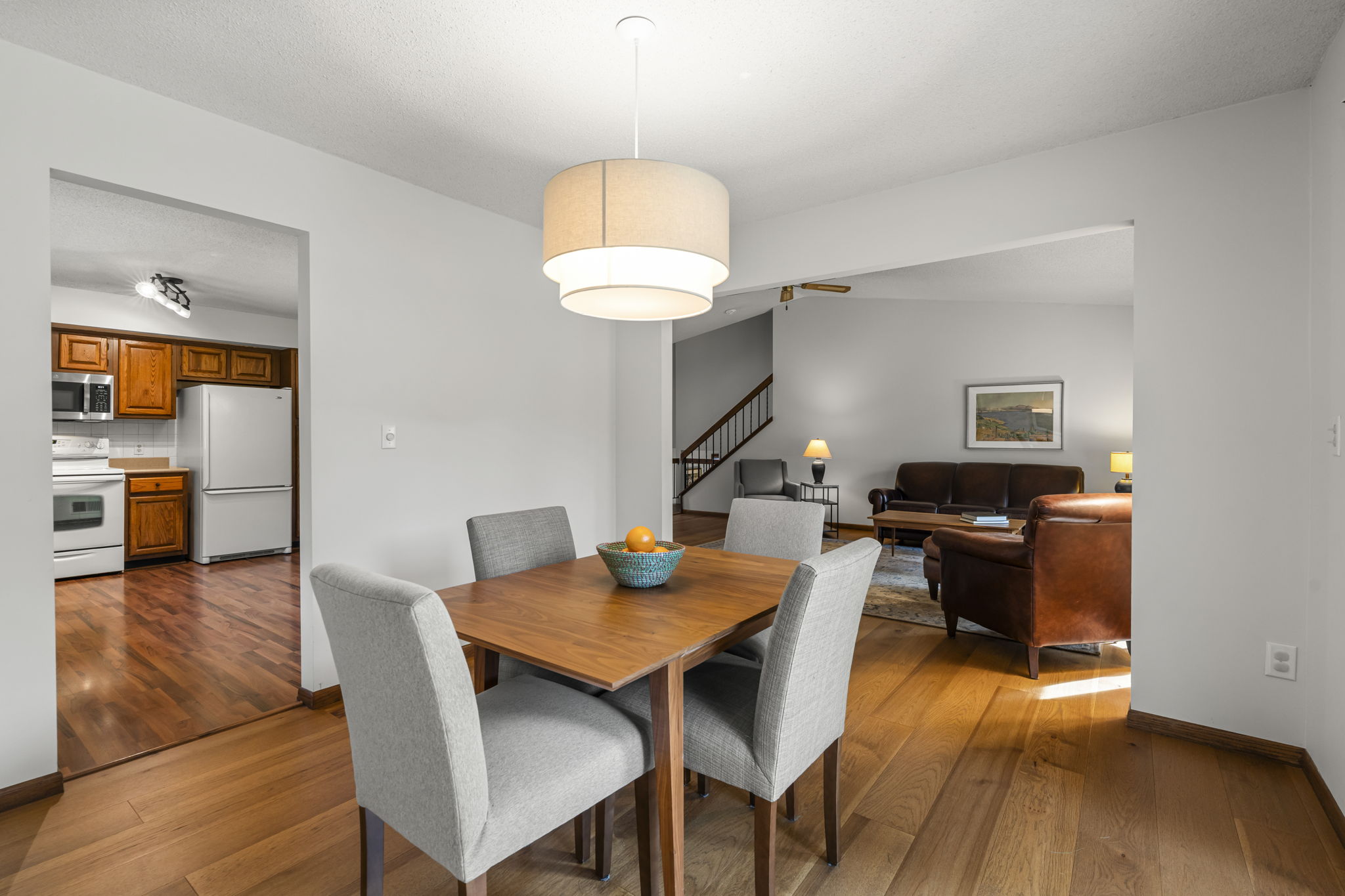 A modern dining area with a wooden table and four gray upholstered chairs, illuminated by a large round ceiling light. Enjoy views into the kitchen and living room in this stylish home for sale by Beekeeper Realty at 9333 Colorado Circle.