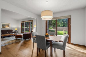 A modern Minnesota dining area with a wooden table, gray chairs, and a pendant light beside a sliding glass door leading to the backyard. A cozy living room with leather armchairs is visible—listed by Beekeeper Realty as a home for sale.
