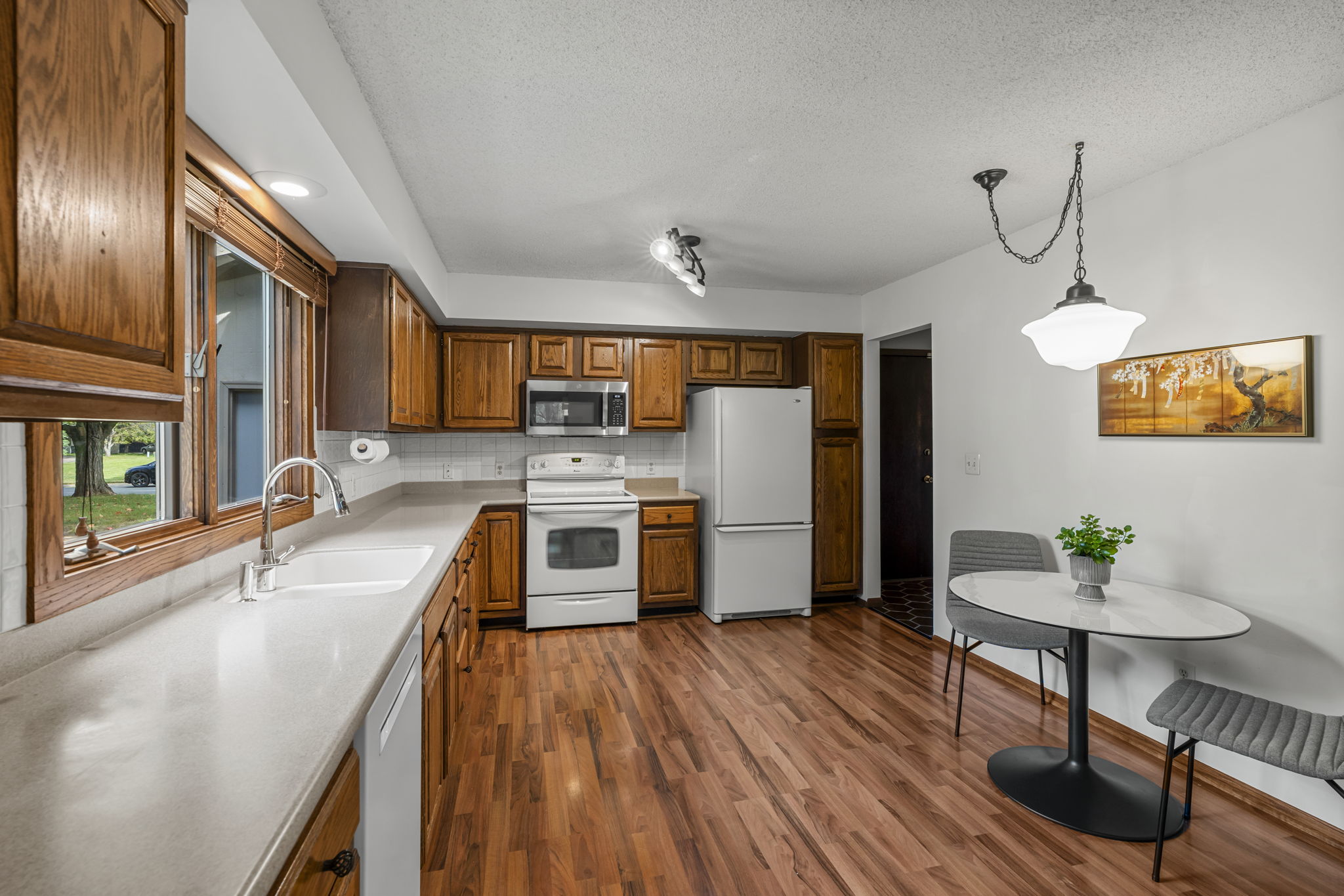 A kitchen with wooden cabinets, white appliances, and a light countertop. A round dining table sits by the window overlooking a green yard. Wood laminate floors complete this cozy Minnesota home for sale from Beekeeper Realty.