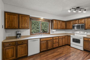 Spacious kitchen in this Bloomington home for sale featuring wooden cabinets, white appliances including a stove, microwave, dishwasher, coffee maker, double sink under a window, wood flooring, and ceiling light fixture. Listed by Beekeeper Realty.