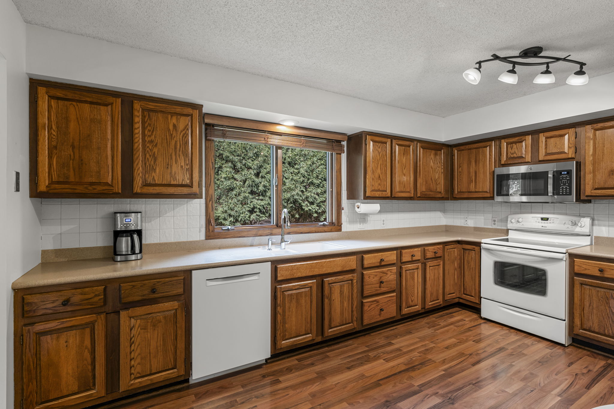 Spacious kitchen in this Bloomington home for sale featuring wooden cabinets, white appliances including a stove, microwave, dishwasher, coffee maker, double sink under a window, wood flooring, and ceiling light fixture. Listed by Beekeeper Realty.