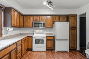 A kitchen in the home for sale at 9333 Colorado Circle features wooden cabinets, white countertops, a white stove and oven, a white refrigerator, a microwave, overhead lighting, wood floors, and a window with blinds to the left. Listed by Beekeeper Realty.