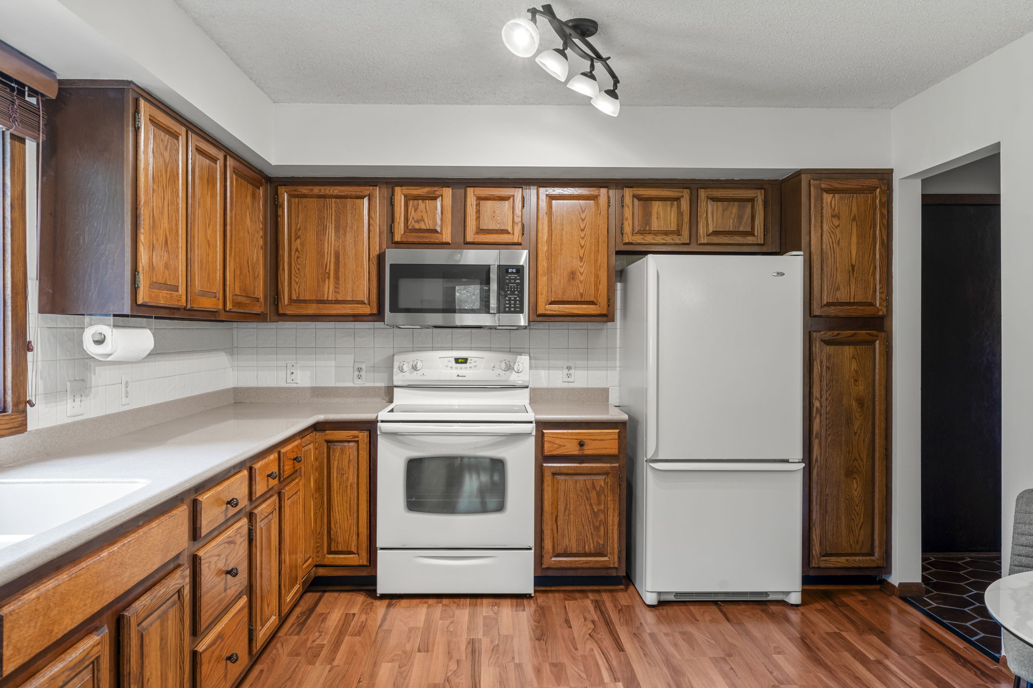 A kitchen in the home for sale at 9333 Colorado Circle features wooden cabinets, white countertops, a white stove and oven, a white refrigerator, a microwave, overhead lighting, wood floors, and a window with blinds to the left. Listed by Beekeeper Realty.