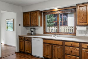 A kitchen at 9333 Colorado Circle features wooden cabinets, a white dishwasher, coffee maker, and a double sink under a large window with blinds and views of trees and a fence. The wood floor flows into the hallway. Listed by Beekeeper Realty in Minnesota.