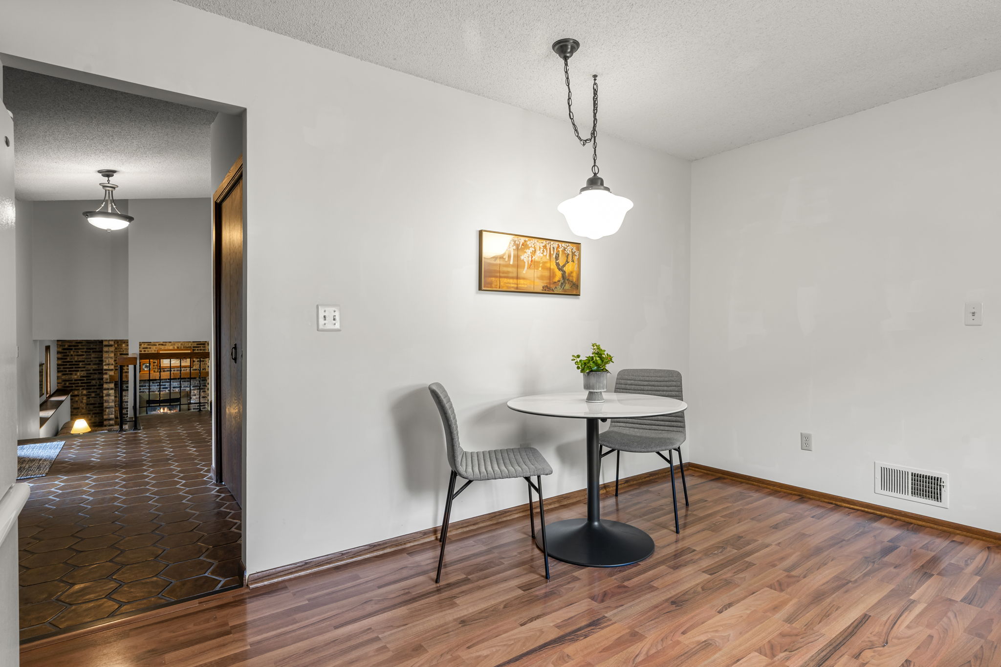 Cozy small dining area in this Bloomington home for sale, featuring two gray chairs and a round table on wood flooring, a pendant light above, framed wall art, and a potted plant; tiled entryway visible in the background.