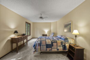 A cozy bedroom in Bloomington, Minnesota with beige walls, carpeted floor, a bed with a colorful patchwork quilt, two nightstands and lamps, a long wooden console table, window views of greenery, and a ceiling fan—listed by Beekeeper Realty.