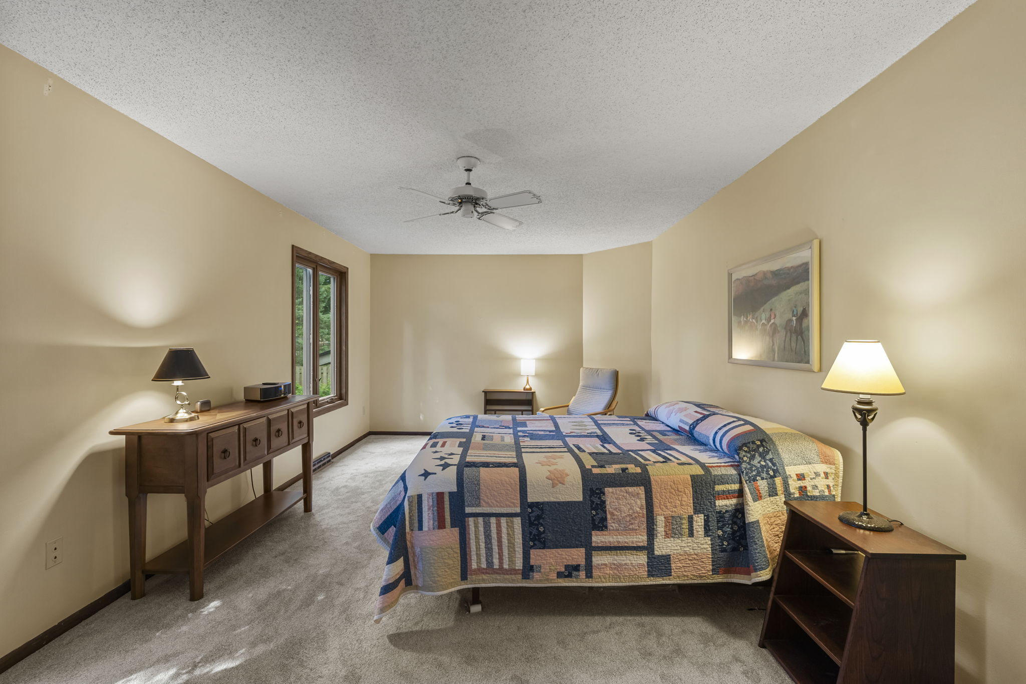 A cozy bedroom in Bloomington, Minnesota with beige walls, carpeted floor, a bed with a colorful patchwork quilt, two nightstands and lamps, a long wooden console table, window views of greenery, and a ceiling fan—listed by Beekeeper Realty.