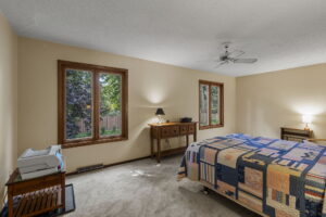 A cozy bedroom in this Minnesota home for sale at 9333 Colorado Circle features beige walls, large windows, a colorful patchwork quilt, a wooden desk with a lamp, and charming views of the green backyard.