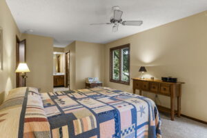 Cozy bedroom at 9333 Colorado Circle in Bloomington, Minnesota, with a patterned quilt, wooden dresser, brown carpet, beige walls, two windows overlooking trees, ceiling fan, and a bathroom visible through an open doorway.