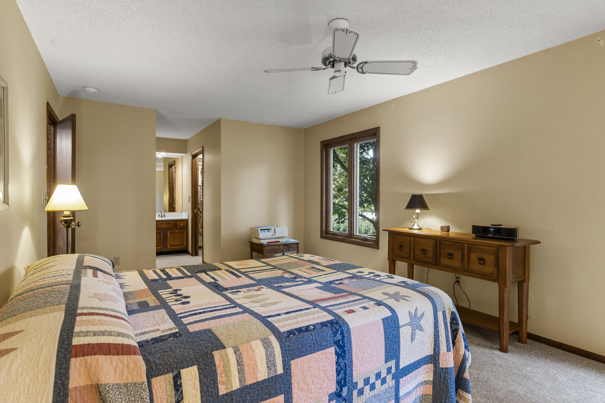 Cozy bedroom at 9333 Colorado Circle in Bloomington, Minnesota, with a patterned quilt, wooden dresser, brown carpet, beige walls, two windows overlooking trees, ceiling fan, and a bathroom visible through an open doorway.