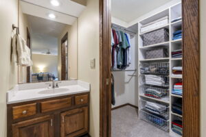 A small bathroom vanity with a sink and mirror sits next to a walk-in closet with shelves holding folded clothes, baskets, and hanging shirts. The room features wood trim, beige walls, and classic Minnesota charm—listed by Beekeeper Realty.