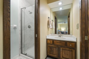 Bathroom with a glass shower on the left, wood vanity with a white countertop and sink in the center, mirror above the sink, and folded towels hanging on the wall. Bedroom visible in the reflection at 9333 Colorado Circle, listed by Beekeeper Realty.
