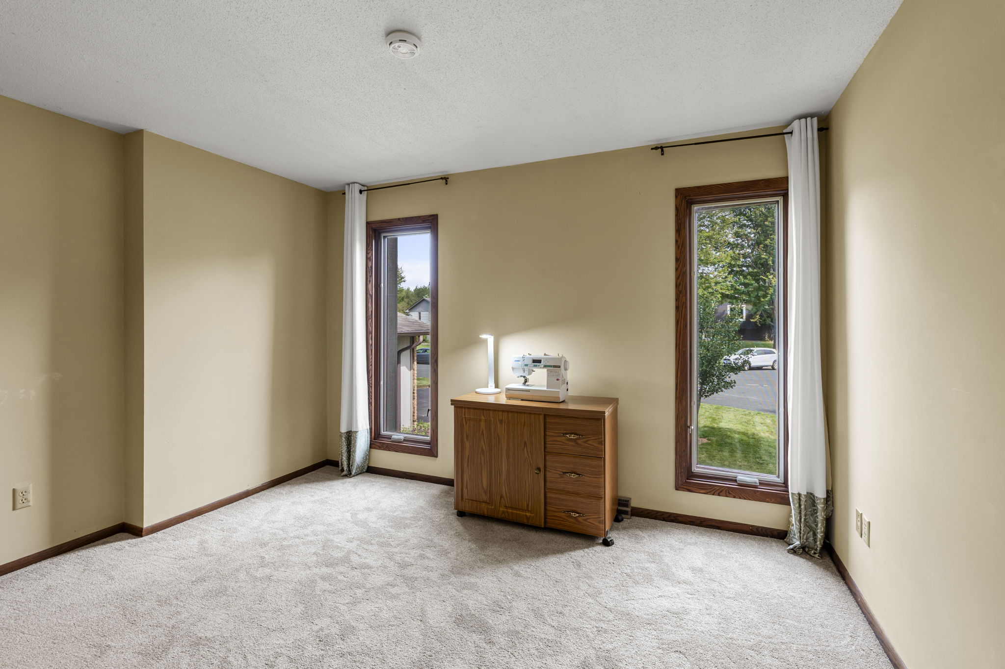 A small room with beige walls and carpet at 9333 Colorado Circle, Bloomington, featuring two tall windows with white curtains and a wooden cabinet with a sewing machine—offered by Beekeeper Realty.