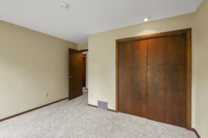 A simple, empty bedroom in Bloomington, Minnesota with beige walls, light carpet flooring, a wooden door, and double-door closet. Presented by Beekeeper Realty. A small vent is near the floor, and a ceiling light sits above the closet.