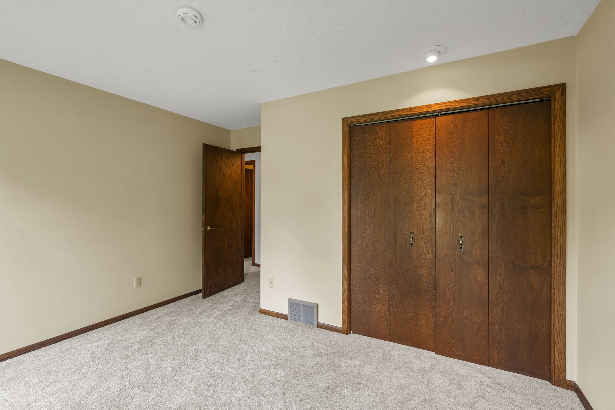 A simple, empty bedroom in Bloomington, Minnesota with beige walls, light carpet flooring, a wooden door, and double-door closet. Presented by Beekeeper Realty. A small vent is near the floor, and a ceiling light sits above the closet.