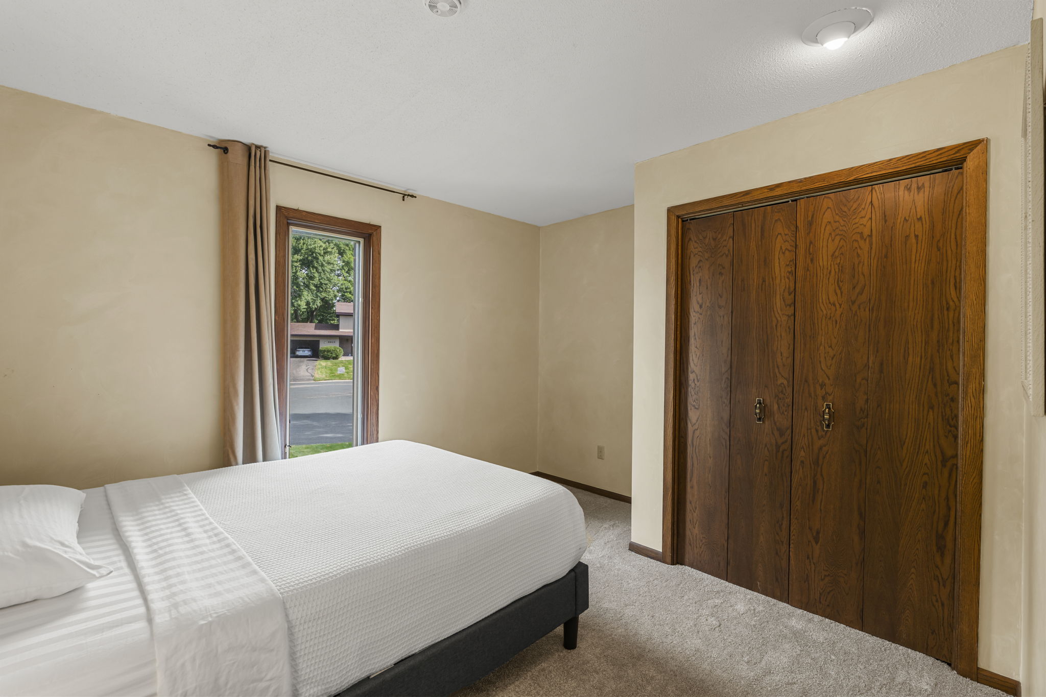 A simple bedroom with beige walls, white bedding, tan curtains framing a view of trees, and a closed wooden closet door—just one of the comfortable spaces in this home for sale at 9333 Colorado Circle by Beekeeper Realty.