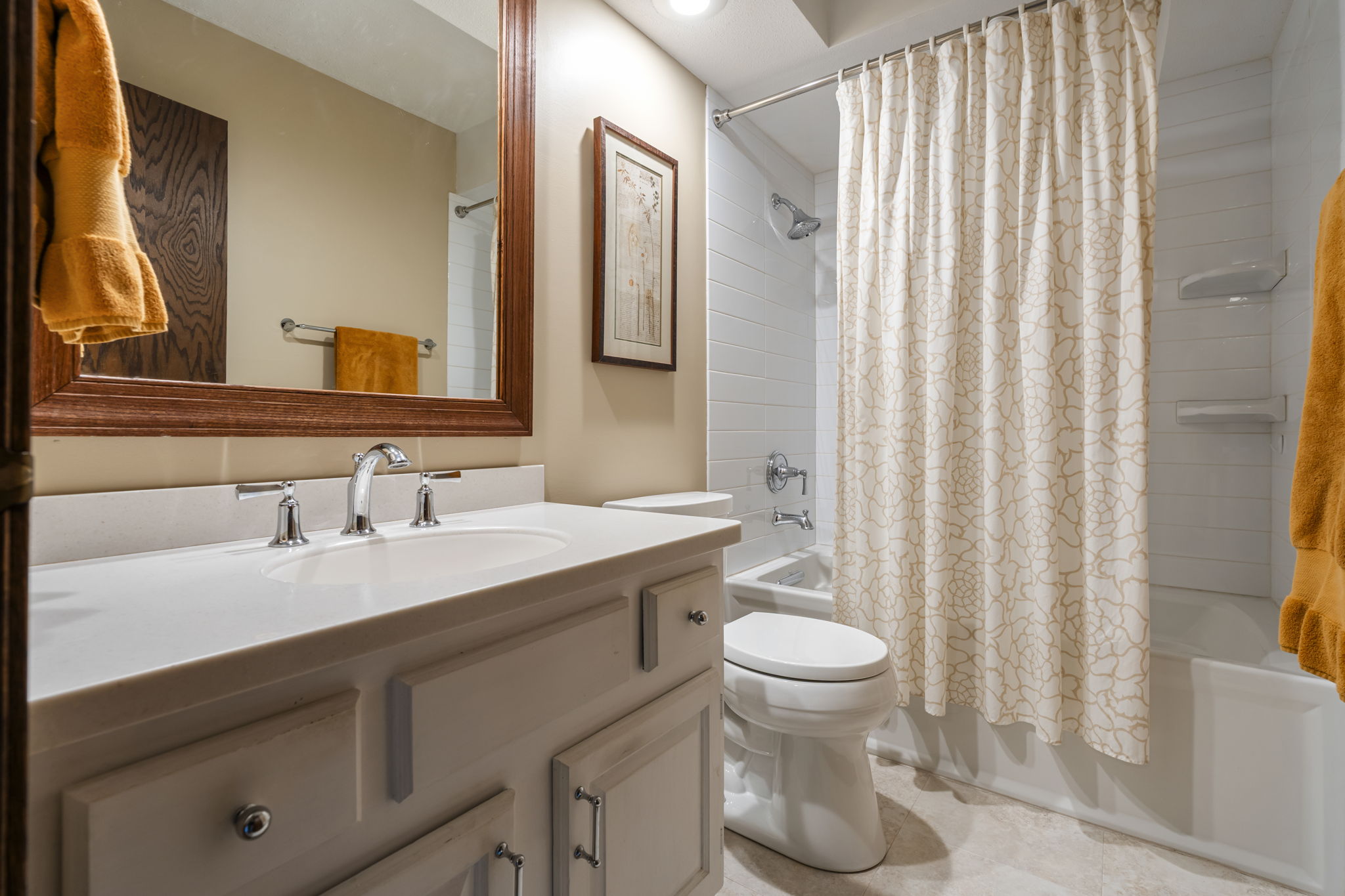 A clean Bloomington bathroom in a Minnesota home for sale features a white vanity and sink, large wooden-framed mirror, toilet, and white bathtub with a patterned shower curtain. Yellow towels hang on the wall, with framed art above the toilet.
