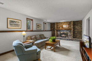 A cozy living room in Minnesota with a brick fireplace, plaid sofa, light blue armchair, wooden coffee table with a plant, side table with a lamp, and framed art on white walls. A TV sits on a stand in the corner of 9333 Colorado Circle.