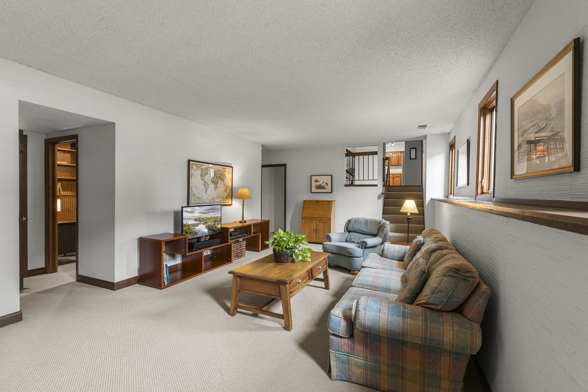 A cozy Bloomington basement living room with a plaid sofa, armchair, wooden coffee table, TV stand, potted plant, wall art, and carpeted floor. Stairs lead up and windows fill this Minnesota home for sale with natural light.