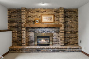 A brick fireplace with a wooden mantel, featuring a framed landscape photo and a small picture on top. The gas insert showcases a visible flame, set in a carpeted room with white walls at 9333 Colorado Circle, listed by Beekeeper Realty in Bloomington.
