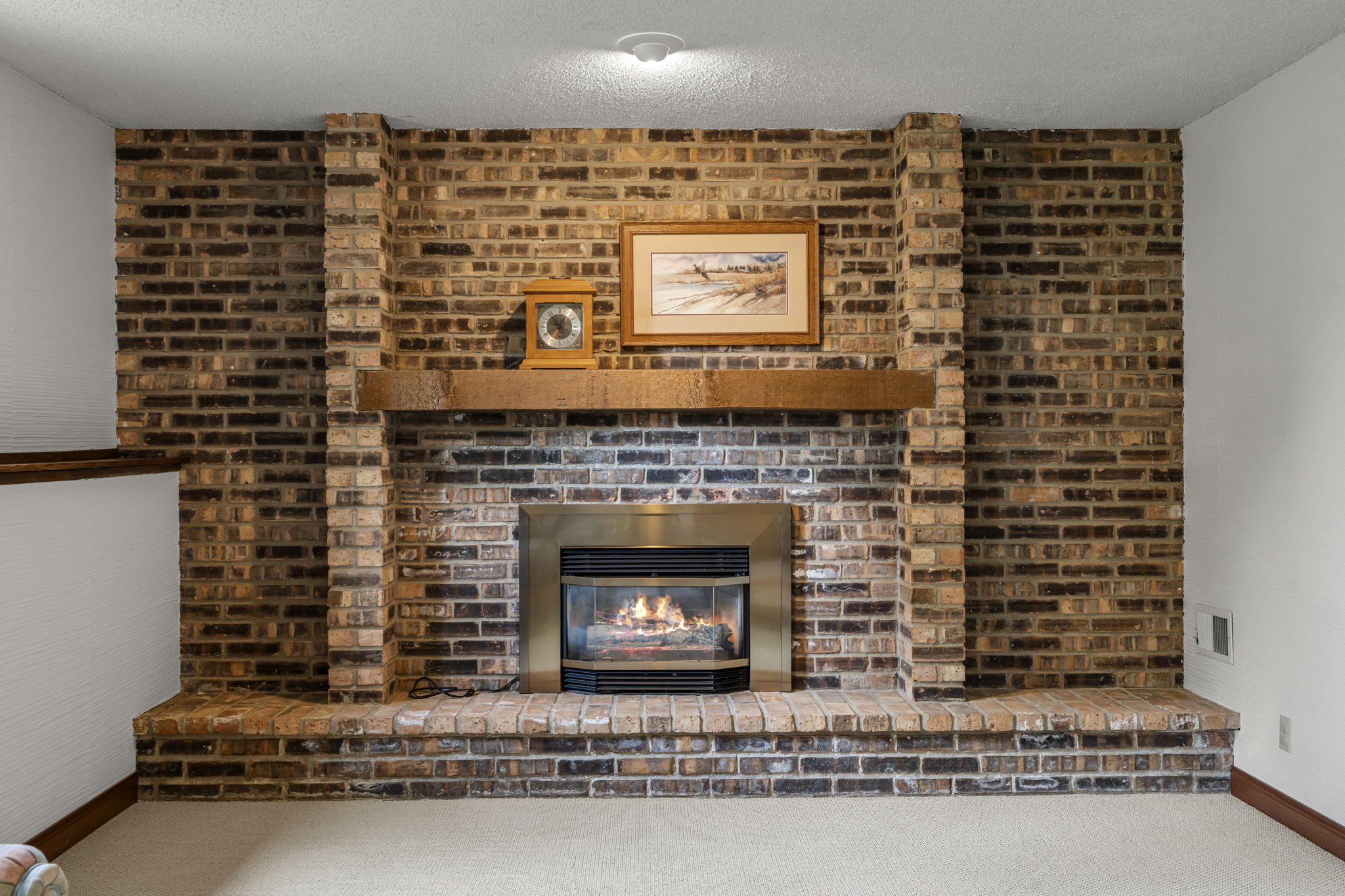 A brick fireplace with a wooden mantel, featuring a framed landscape photo and a small picture on top. The gas insert showcases a visible flame, set in a carpeted room with white walls at 9333 Colorado Circle, listed by Beekeeper Realty in Bloomington.