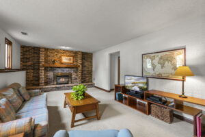 A cozy living room at 9333 Colorado Circle with a plaid sofa, wooden coffee table, TV console, lamp, and map on the wall. The brick fireplace features a framed picture and a potted plant—your next home for sale from Beekeeper Realty.