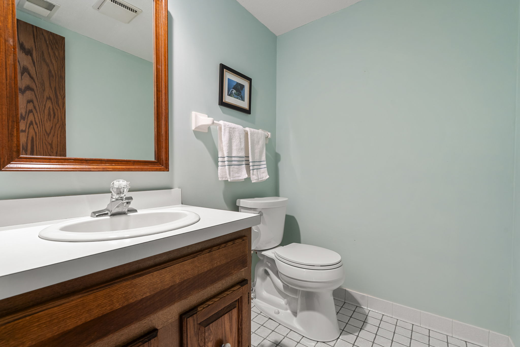 A small bathroom in this Minnesota home for sale features a wooden vanity, white sink, toilet, neatly folded towels on a rack, pale blue walls, a framed picture, and white tile flooring.