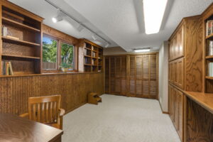 A home office or study room in this Bloomington, Minnesota home for sale features wood-paneled walls, built-in wooden bookshelves, a wooden desk and chair, beige carpet, large windows, and double wooden closet doors.