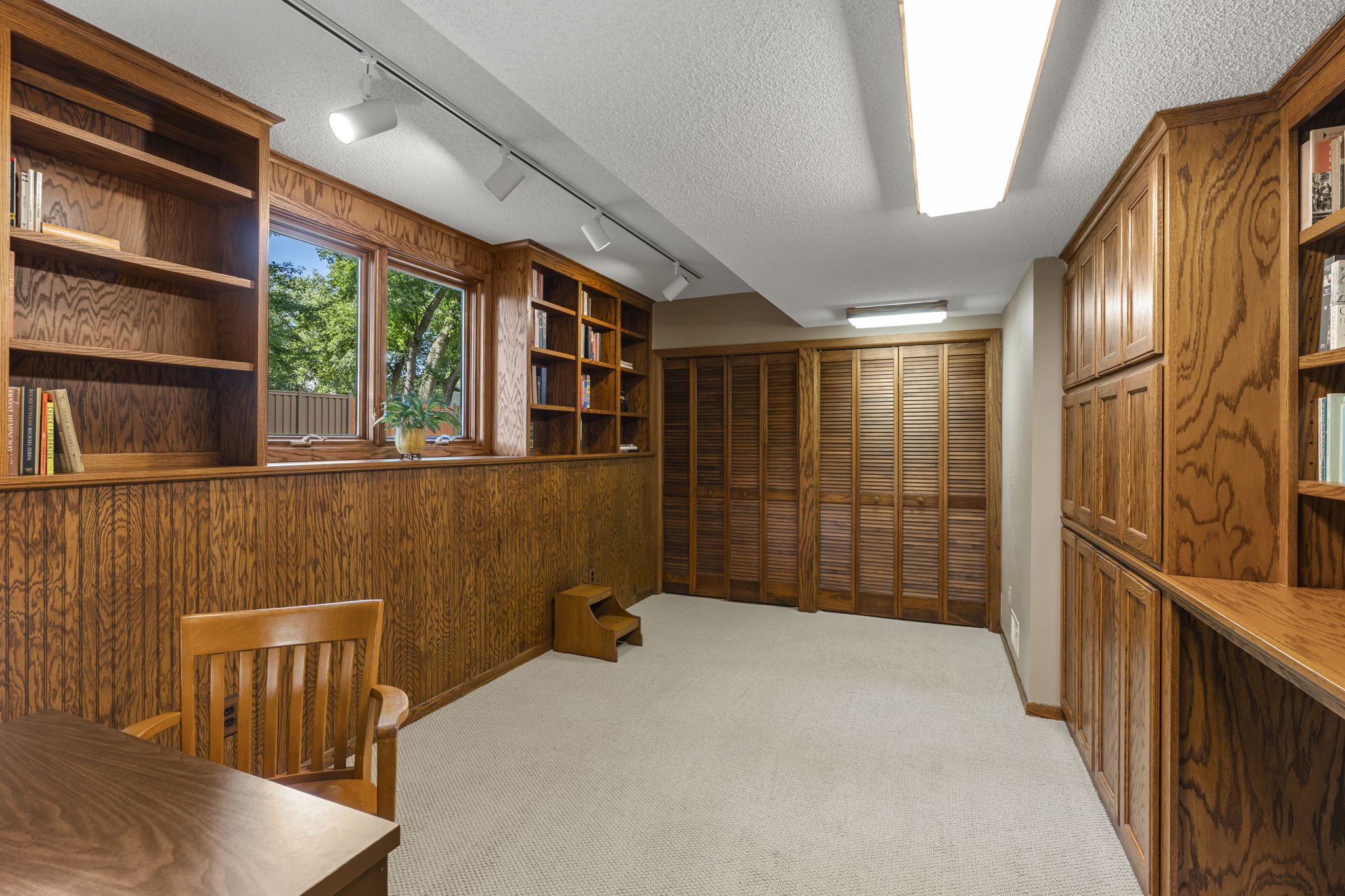 A home office or study room in this Bloomington, Minnesota home for sale features wood-paneled walls, built-in wooden bookshelves, a wooden desk and chair, beige carpet, large windows, and double wooden closet doors.