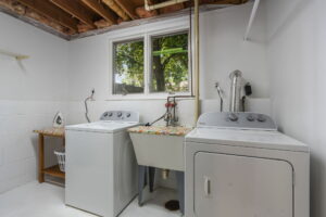 A basement laundry room in this Bloomington, Minnesota home for sale features exposed ceiling beams, a washer and dryer by a utility sink, a small shelf with an iron and laundry basket, and a window overlooking green trees.