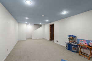 A spacious, carpeted basement room in Bloomington, Minnesota, with white walls, blue ceiling, recessed lighting, and a children’s play kitchen set in the corner near a wooden door. Stairs are visible in the background of 9333 Colorado Circle.