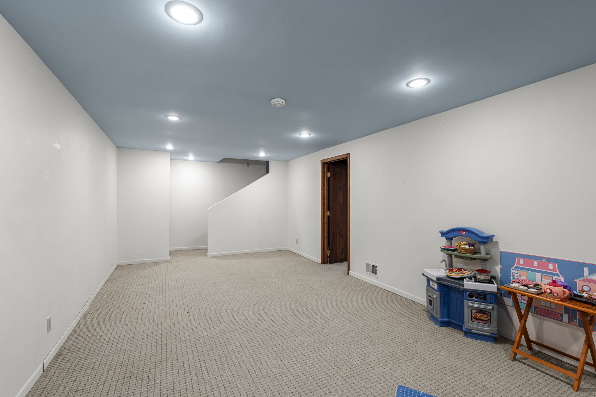 A spacious, carpeted basement room in Bloomington, Minnesota, with white walls, blue ceiling, recessed lighting, and a children’s play kitchen set in the corner near a wooden door. Stairs are visible in the background of 9333 Colorado Circle.