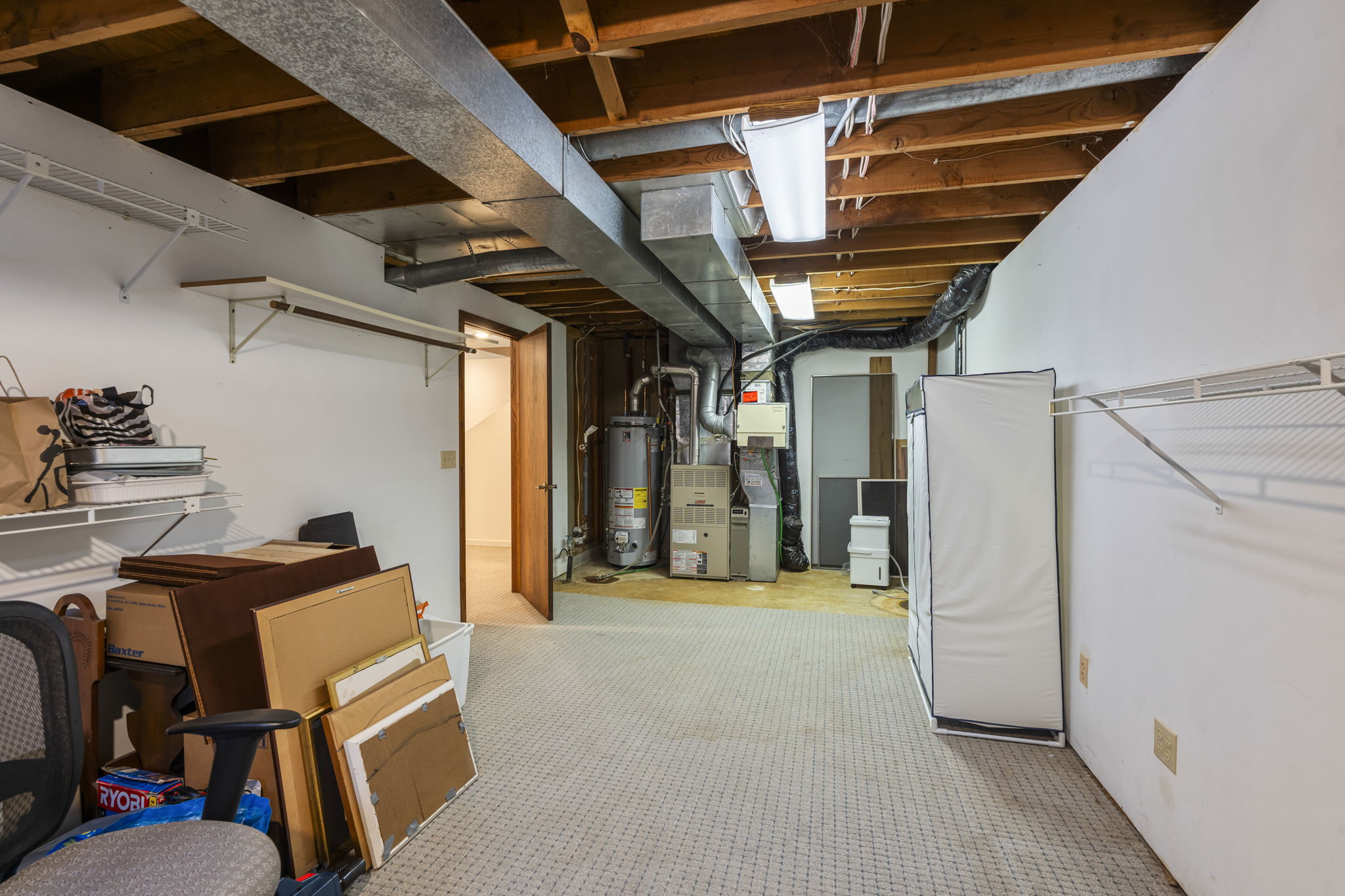 Unfinished basement at 9333 Colorado Circle in Bloomington features exposed ceiling beams, water heater, HVAC unit, storage shelves, wire racks, small desk with chair, and various stored items. Well-lit with fluorescent lighting—listed by Beekeeper Realty.