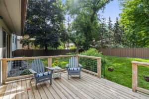 A wooden deck with two gray Adirondack chairs and a small glass table overlooks a lush, green backyard bordered by a brown fence and tall trees—perfect for relaxing at this Minnesota home for sale by Beekeeper Realty.