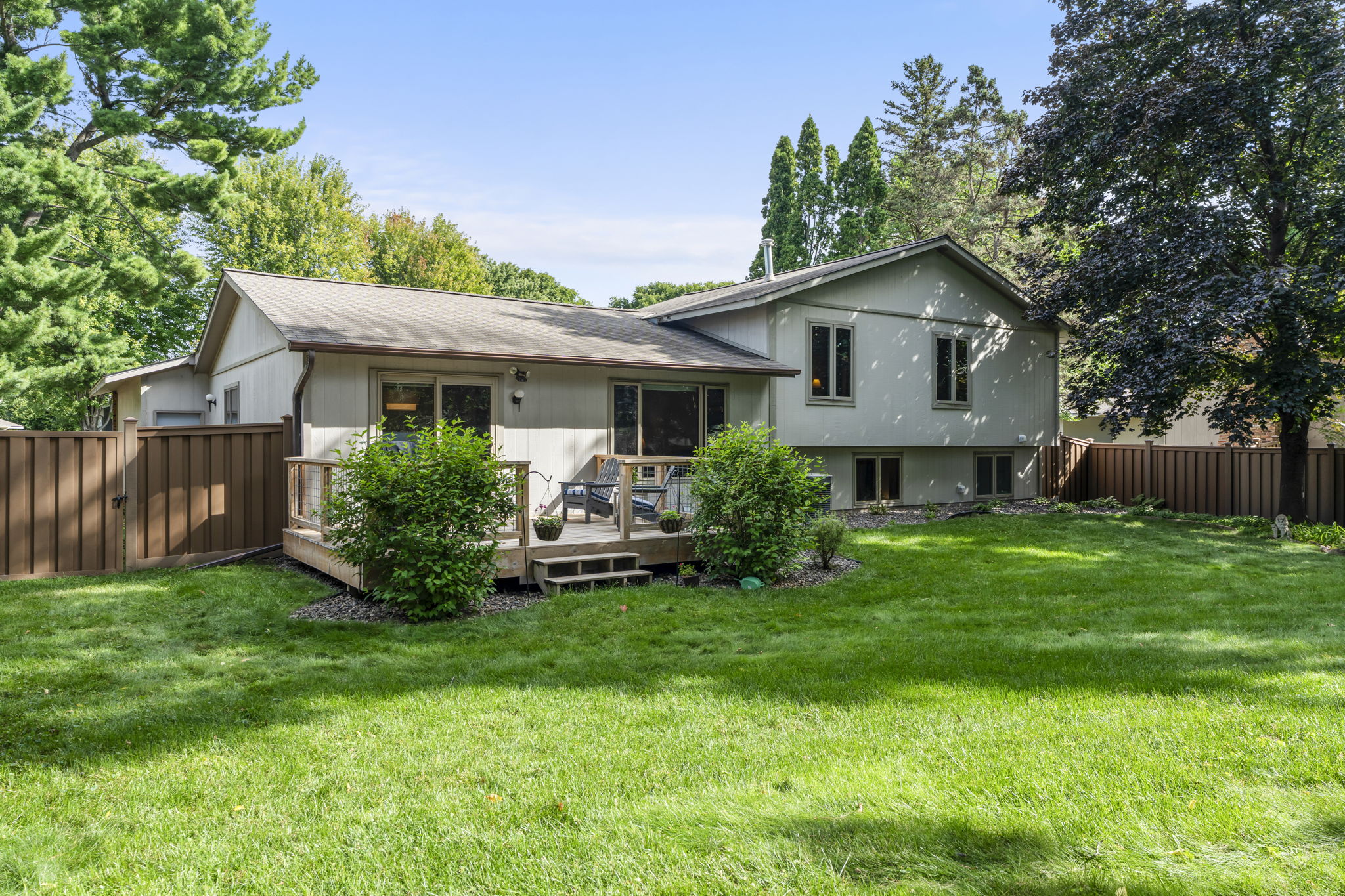 A light gray split-level home for sale by Beekeeper Realty, 9333 Colorado Circle, featuring a wooden deck, patio chairs, green bushes, and a fenced backyard with lush grass and mature trees.