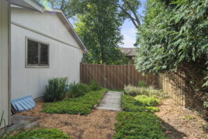 A small backyard at 9333 Colorado Circle features a wood-chip path bordered by green shrubs, a beige fence, and a white house on the left; tall trees provide shade in the background—discover this Bloomington home for sale.