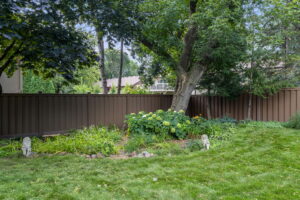 The backyard at 9333 Colorado Circle in Minnesota features lush green grass, a large tree, leafy plants, yellow flowers, and two small stone animal statues along a brown wooden fence, with houses and more trees visible beyond.