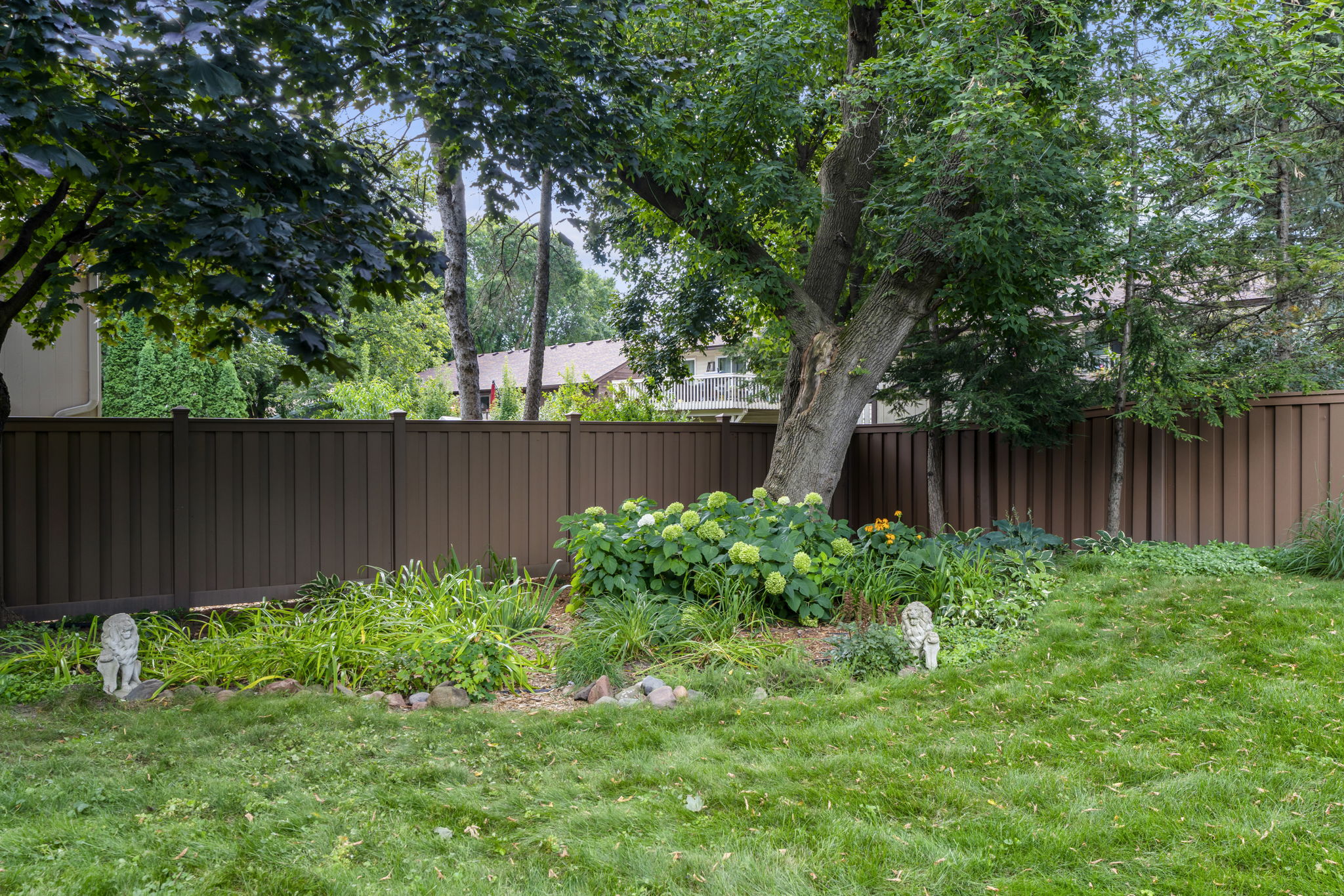 The backyard at 9333 Colorado Circle in Minnesota features lush green grass, a large tree, leafy plants, yellow flowers, and two small stone animal statues along a brown wooden fence, with houses and more trees visible beyond.