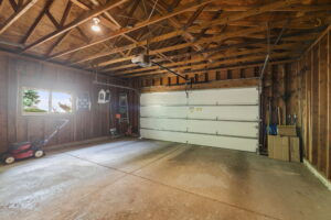 A spacious, empty two-car garage in Bloomington at 9333 Colorado Circle features exposed wooden beams, a white garage door, a window, and garden tools, a lawnmower, and boxes neatly stored along the walls.