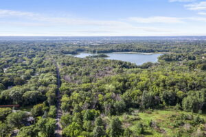Aerial view of a lush, green forest with a winding road leading to 9333 Colorado Circle, a beautiful home for sale near a large lake, with the sprawling Bloomington cityscape visible on the distant horizon under a blue sky.