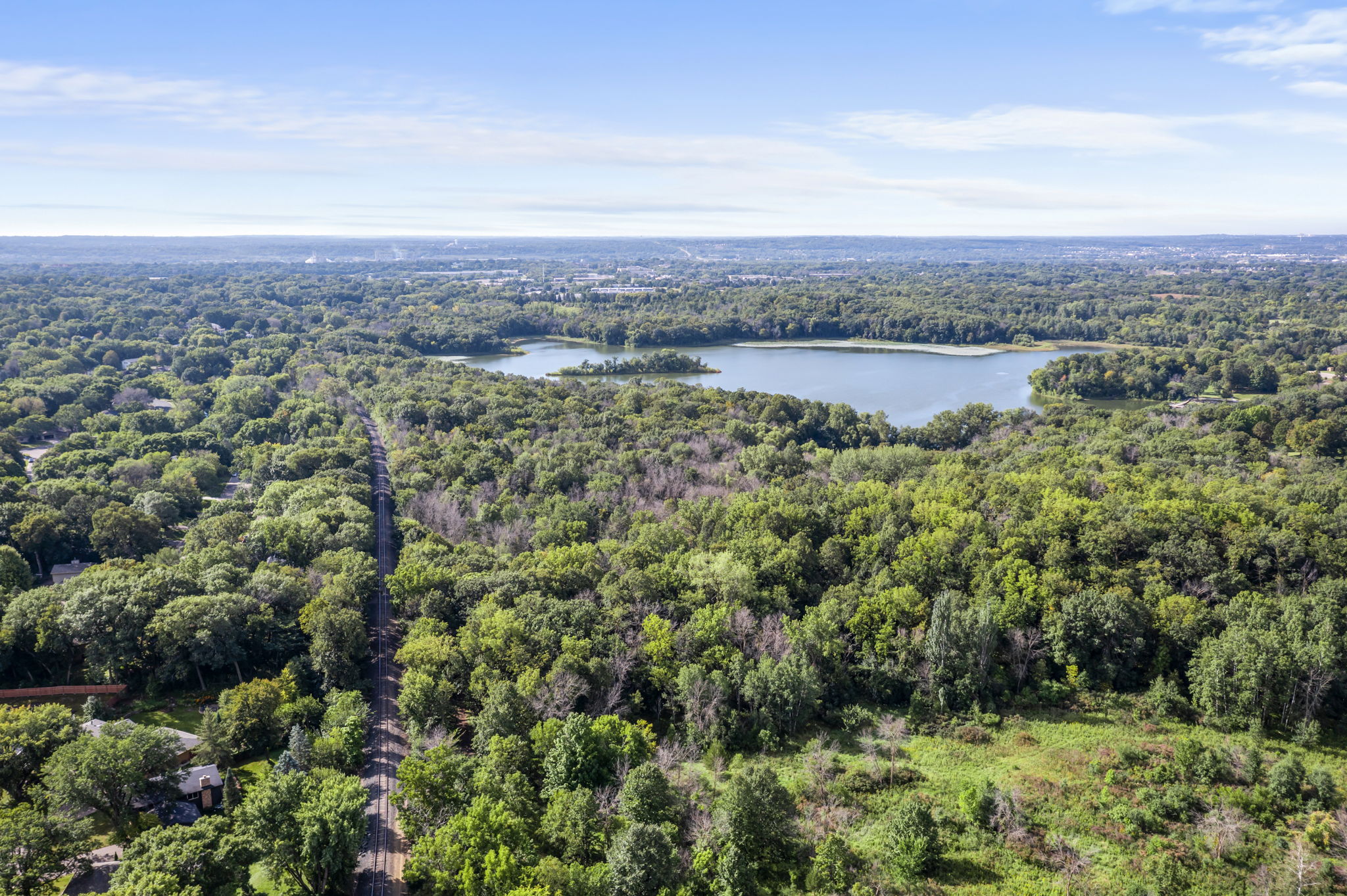 Aerial view of a lush, green forest with a winding road leading to 9333 Colorado Circle, a beautiful home for sale near a large lake, with the sprawling Bloomington cityscape visible on the distant horizon under a blue sky.