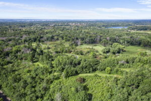Aerial view of a lush green forest near 9333 Colorado Circle, Minnesota, with winding trails, open grassy areas, and a distant body of water under a blue sky with light clouds—perfect setting for your next home for sale.