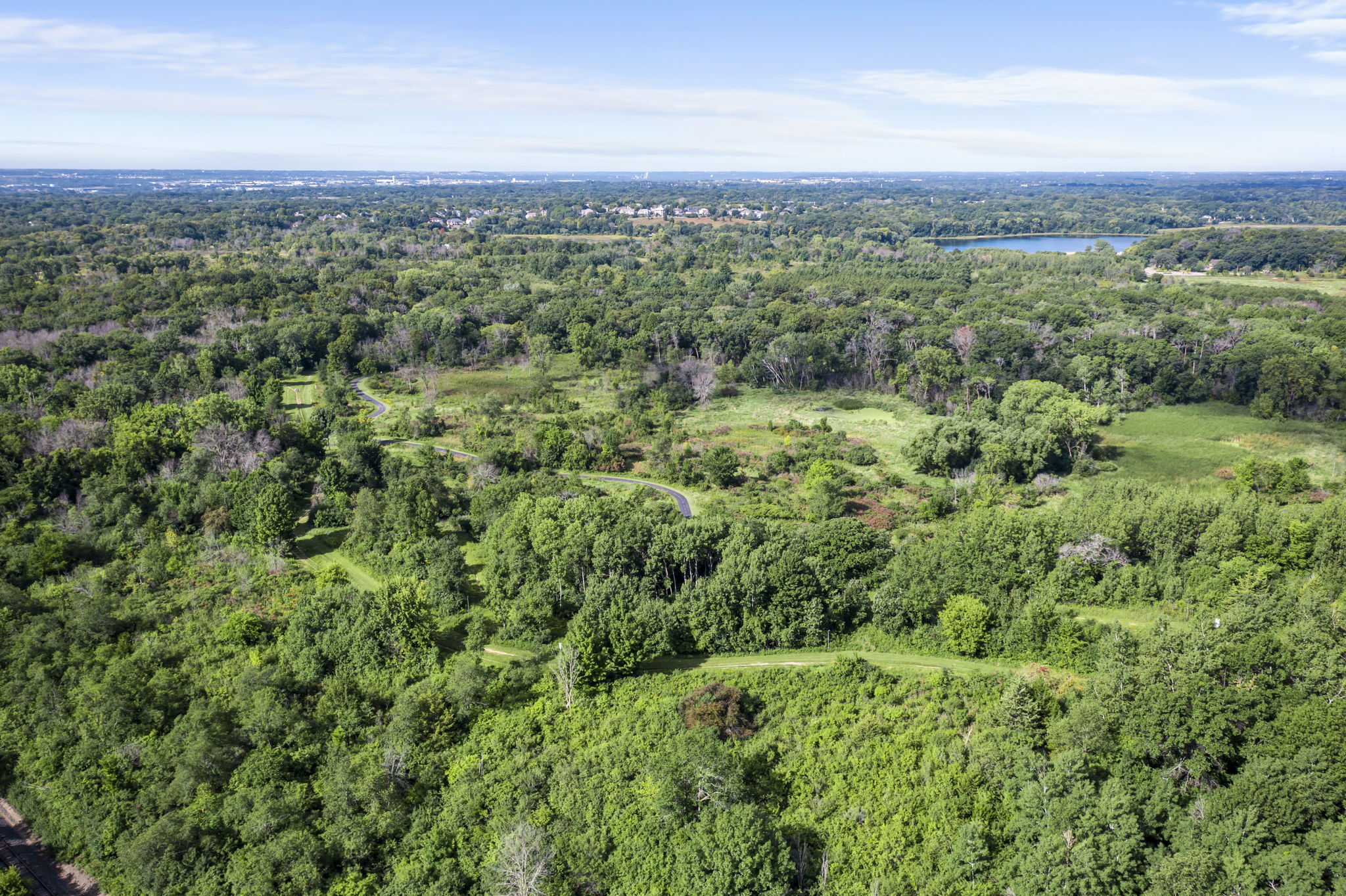 Aerial view of a lush green forest near 9333 Colorado Circle, Minnesota, with winding trails, open grassy areas, and a distant body of water under a blue sky with light clouds—perfect setting for your next home for sale.